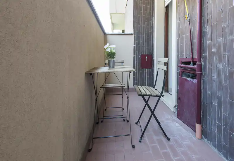 Piccolo balcone arredato con un tavolino bistro e due sedie pieghevoli, vaso di fiori e pavimento piastrellato — perfetto per il caffè mattutino.