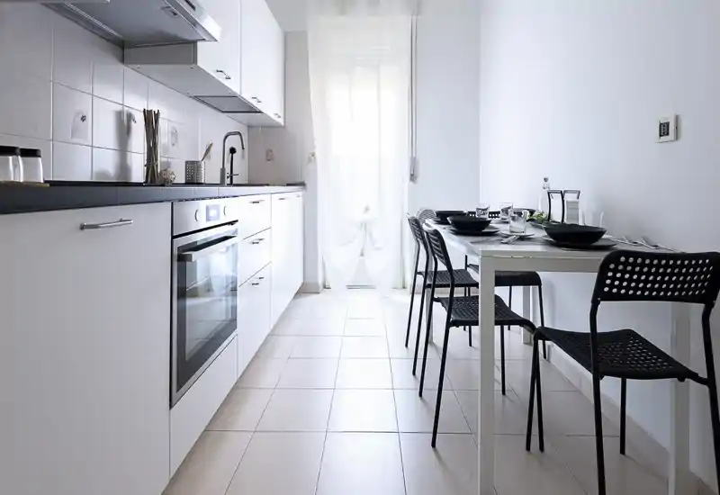 Cucina moderna a corridoio, luminosa, con mobili bianchi, forno integrato e un piccolo tavolo da pranzo per quattro. Stile pulito e minimal con luce naturale che rende l’ambiente arioso.