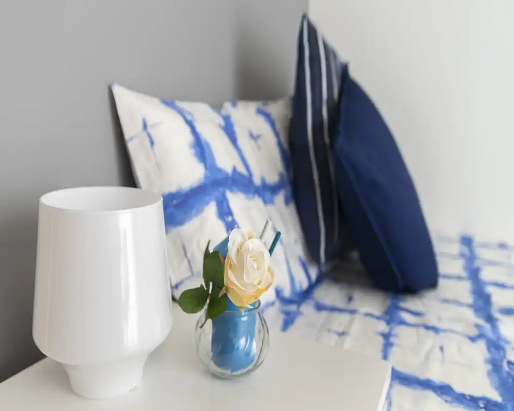 Rincón de dormitorio luminoso y cuidado con ropa de cama azul y blanca estampada, cojines y mesita con lámpara y jarrón decorativo—limpio, fresco y acogedor.