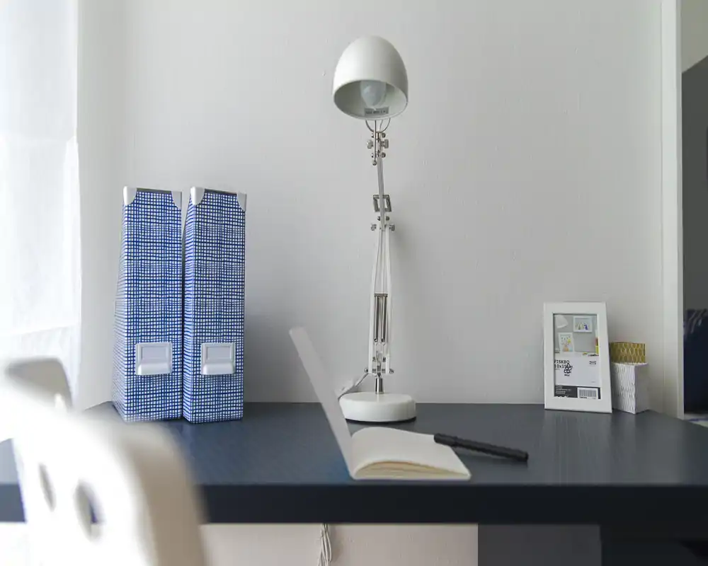 Rincón de escritorio luminoso y minimalista con una lámpara blanca, dos archivadores azules y un cuaderno abierto — ideal como espacio de trabajo o estudio compacto.