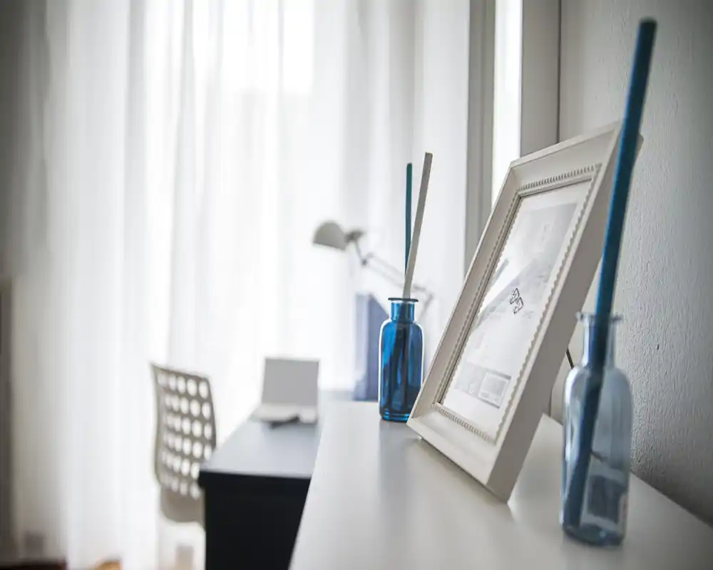 Detalle luminoso de una sala de estar minimalista con botellas de vidrio azul y un marco en una consola, luz natural suave que atraviesa cortinas translúcidas y un escritorio/silla en el fondo.