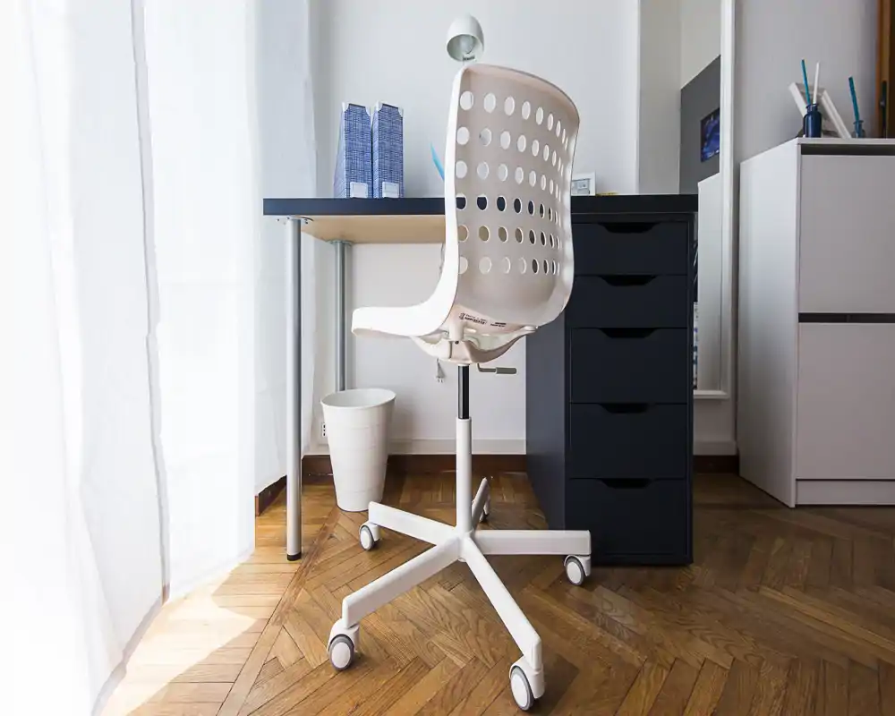 Rincón de oficina en casa luminoso y moderno con silla giratoria blanca, escritorio sencillo y cajonera junto a una gran ventana que aporta luz natural.