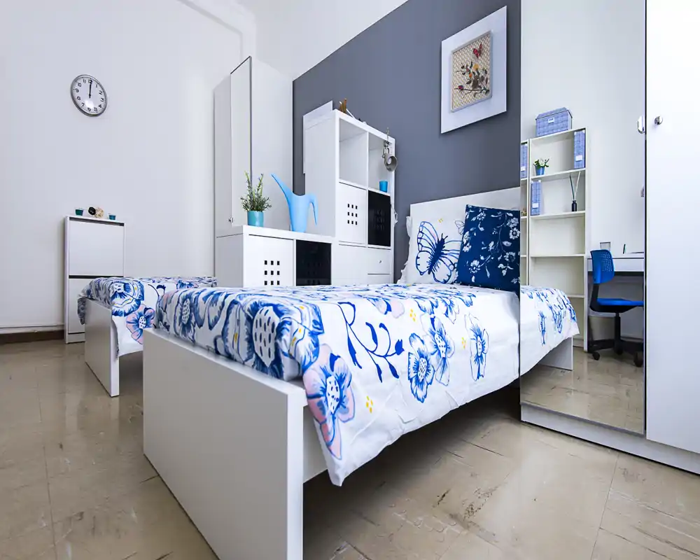 Camera da letto luminosa e moderna con letto bianco e biancheria floreale blu, scaffalature coordinate e una scrivania — ideale per studenti o ospiti.