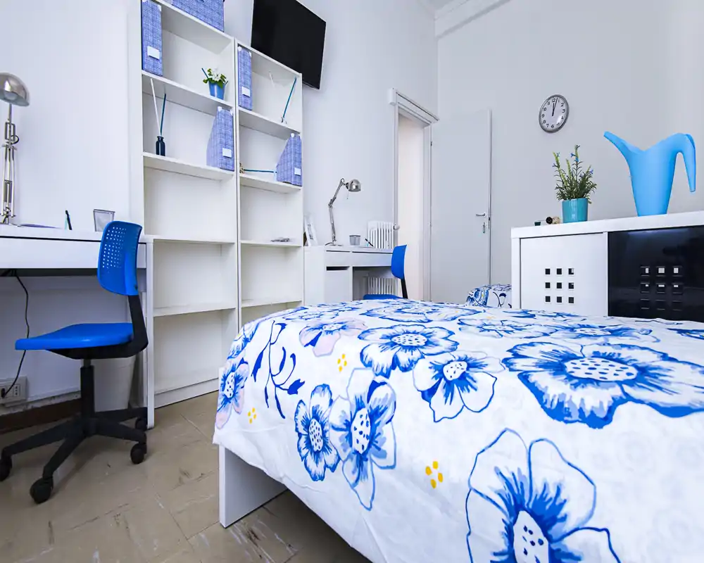 Camera da letto luminosa e ordinata con copriletto blu e bianco, scrivania e scaffalatura. La stanza è ben illuminata e allestita in modo curato, ideale per annunci di affitto.