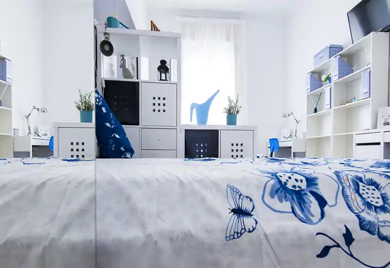 Bright, modern bedroom with a blue-and-white floral duvet, shelving units and natural light from a central window — a clean, welcoming space ideal for a rental listing.