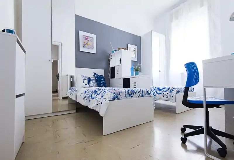 Bright, modern bedroom with two single beds featuring blue floral bedding, white furniture and a large window providing abundant natural light.