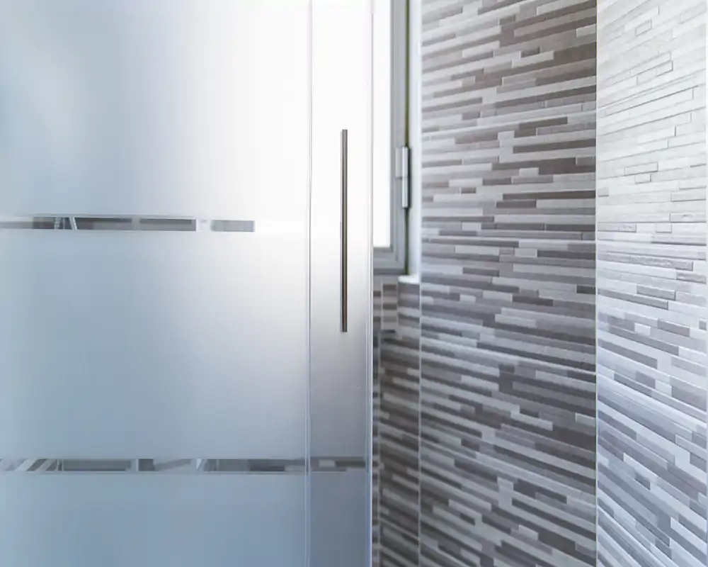 Primo piano di una doccia moderna con porta in vetro satinato scorrevole e maniglia in metallo, su parete piastrellata con mosaico orizzontale e luce naturale dalla finestra.