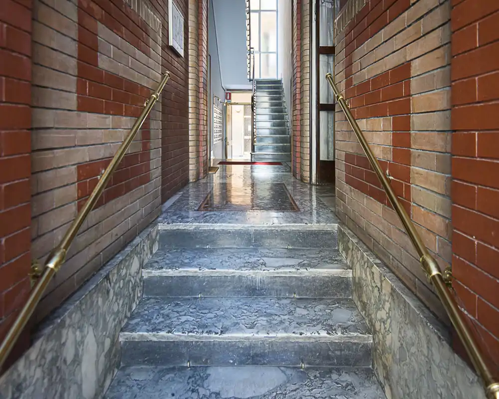 Ingresso dell'edificio luminoso e pulito con gradini in marmo, corrimano in ottone e pareti in mattoni rossi che conducono a una scala interna.