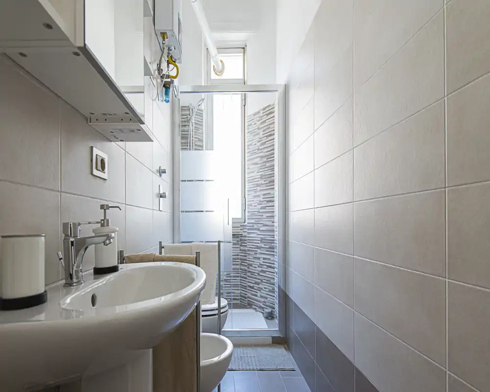 Bagno moderno e luminoso con lavabo, bidet, WC e una doccia con cabina in vetro sul fondo; piastrelle neutre e sanitari puliti creano un ambiente curato.