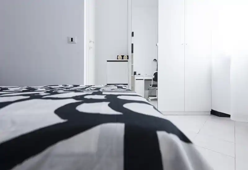 Minimal, bright bedroom view focused on a patterned bedspread with a white wardrobe and desk area in the background. The photo emphasizes cleanliness, natural light and modern minimalist styling.