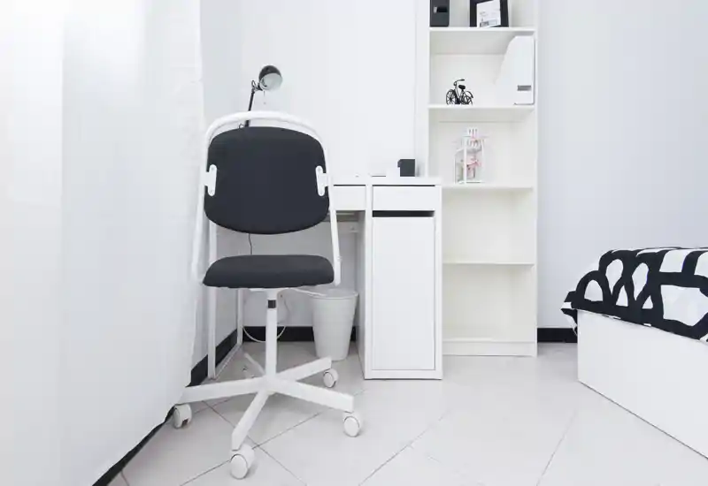 Minimal, bright bedroom corner with a white desk, office chair and a partially visible bed with black-and-white bedding. The space is neat, well-lit and furnished in a clean, modern minimalist style.