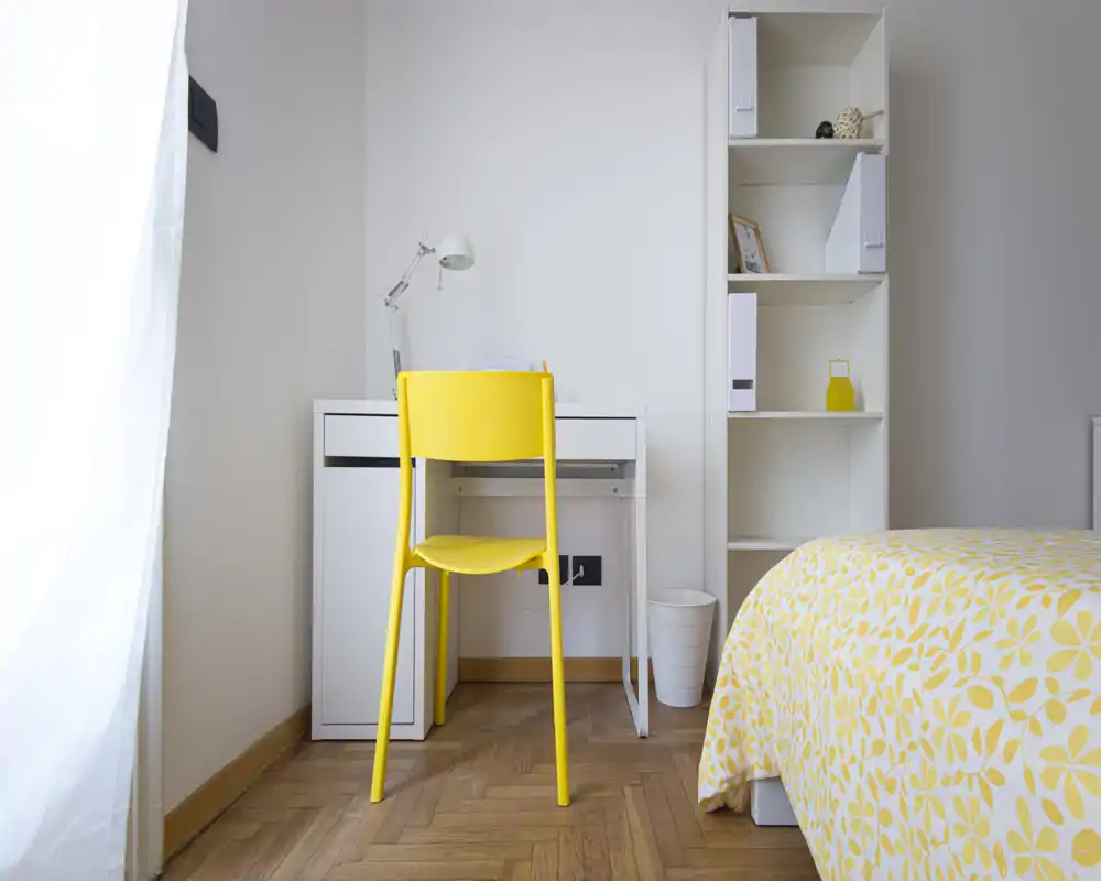 Rincón de dormitorio luminoso y moderno con escritorio blanco, silla amarilla y colcha estampada en amarillo. Líneas limpias y luz natural crean un acogedor espacio de trabajo junto a la cama.