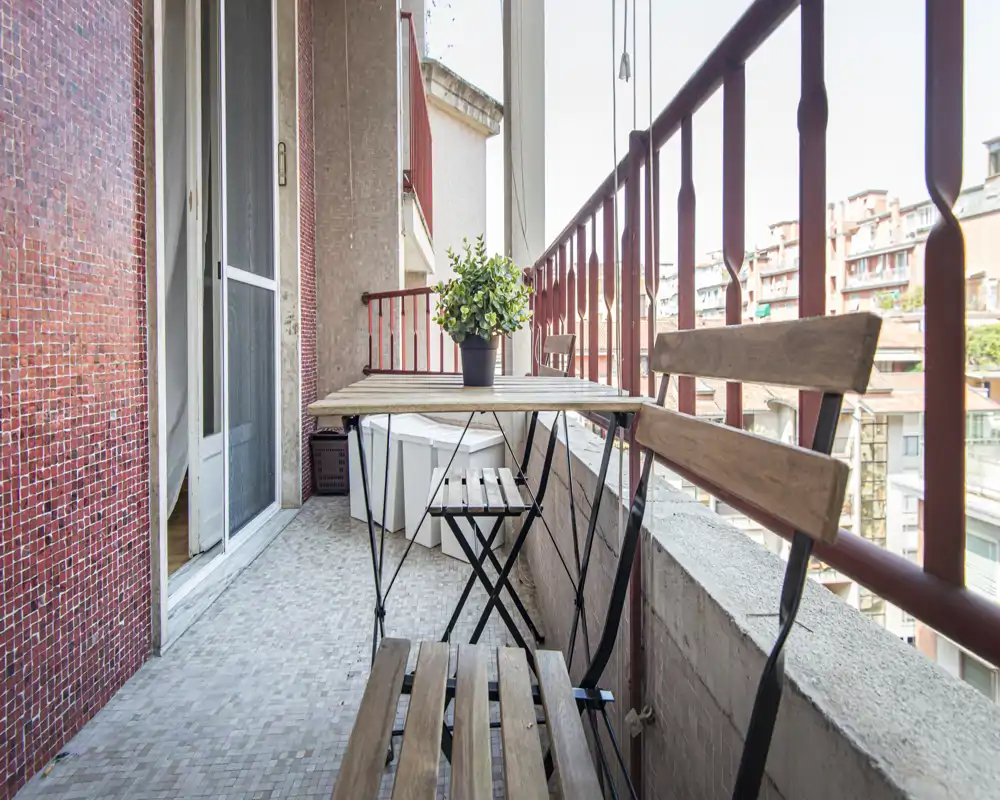 Balcón estrecho y soleado con pequeña mesa y sillas de estilo bistró en madera, suelo de baldosas y una planta en maceta — ideal para el café de la mañana con vistas a la ciudad.