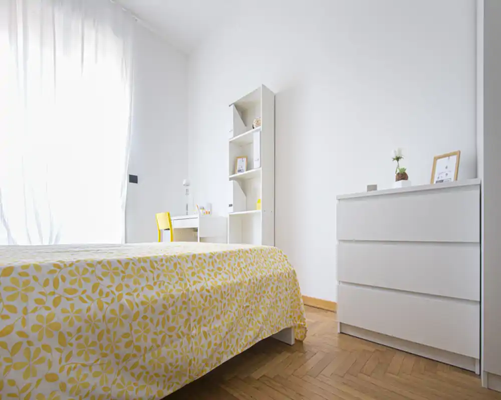 Dormitorio luminoso y minimalista con colcha amarilla de estampado, muebles blancos y un pequeño escritorio junto a una gran ventana que aporta abundante luz natural.