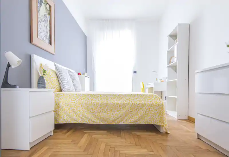 Dormitorio moderno y luminoso con cama doble vestida con funda nórdica amarilla estampada, mesitas de noche y área de escritorio junto a una gran ventana. Suelo de parqué y muebles blancos aportan una atmósfera fresca y acogedora ideal para anuncios.