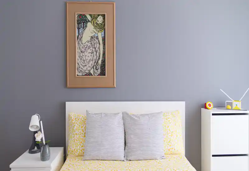 Dormitorio moderno y luminoso con ropa de cama amarilla estampada, cojines a rayas y una obra enmarcada sobre la cabecera blanca. Mesita minimalista con lámpara y planta que crea un ambiente limpio y acogedor.