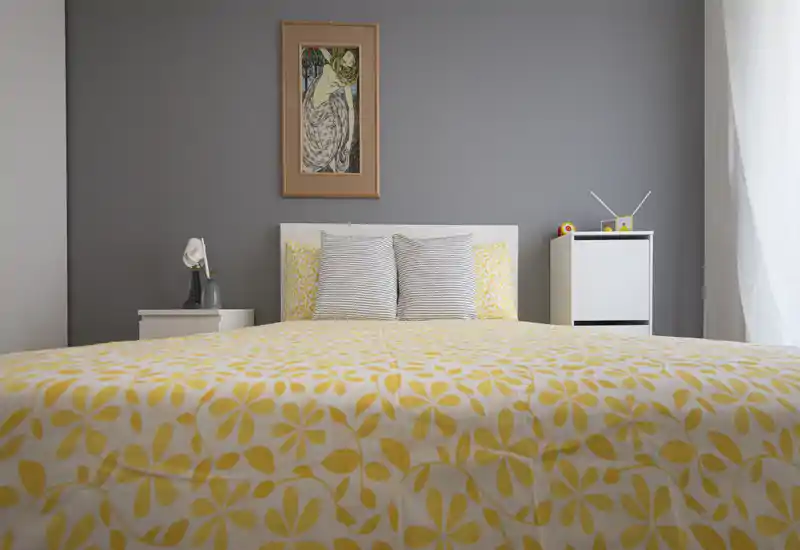 Dormitorio moderno y luminoso con una cama grande vestida con edredón amarillo floral, pared gris neutra y mesillas sencillas — aireado y bien presentado para alquileres.