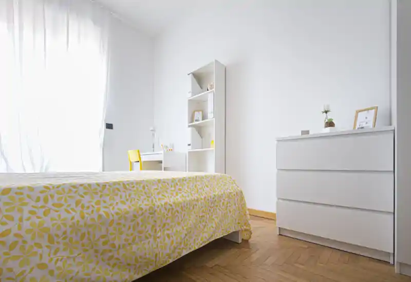 Dormitorio luminoso y minimalista con colcha amarilla de estampado, muebles blancos y un pequeño escritorio junto a una gran ventana que aporta abundante luz natural.