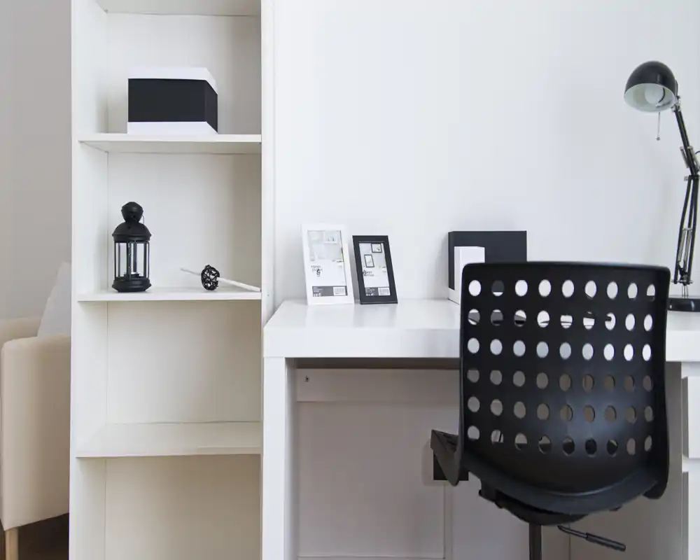 A minimal, well-lit living/work corner with a white desk, shelving and a black perforated office chair — neat and functional for remote work.