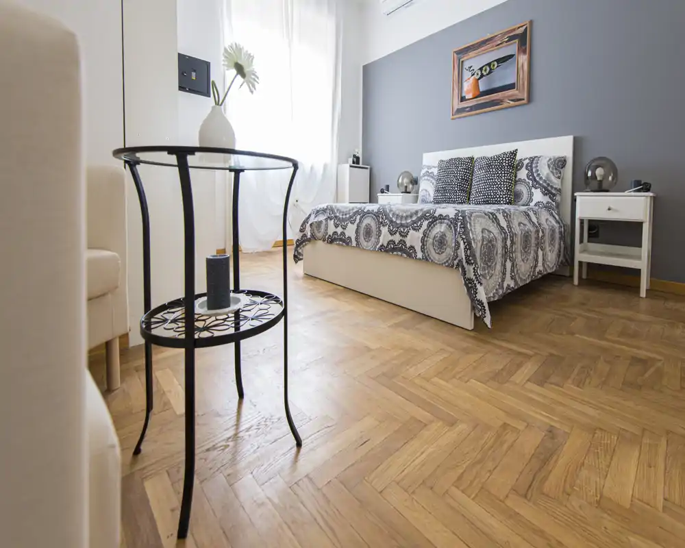 Bright, well-staged bedroom with a double bed dressed in patterned linens, bedside tables with lamps, and natural light from a large window. Hardwood herringbone floor and neutral decor create a calm, modern atmosphere ideal for rental listings.