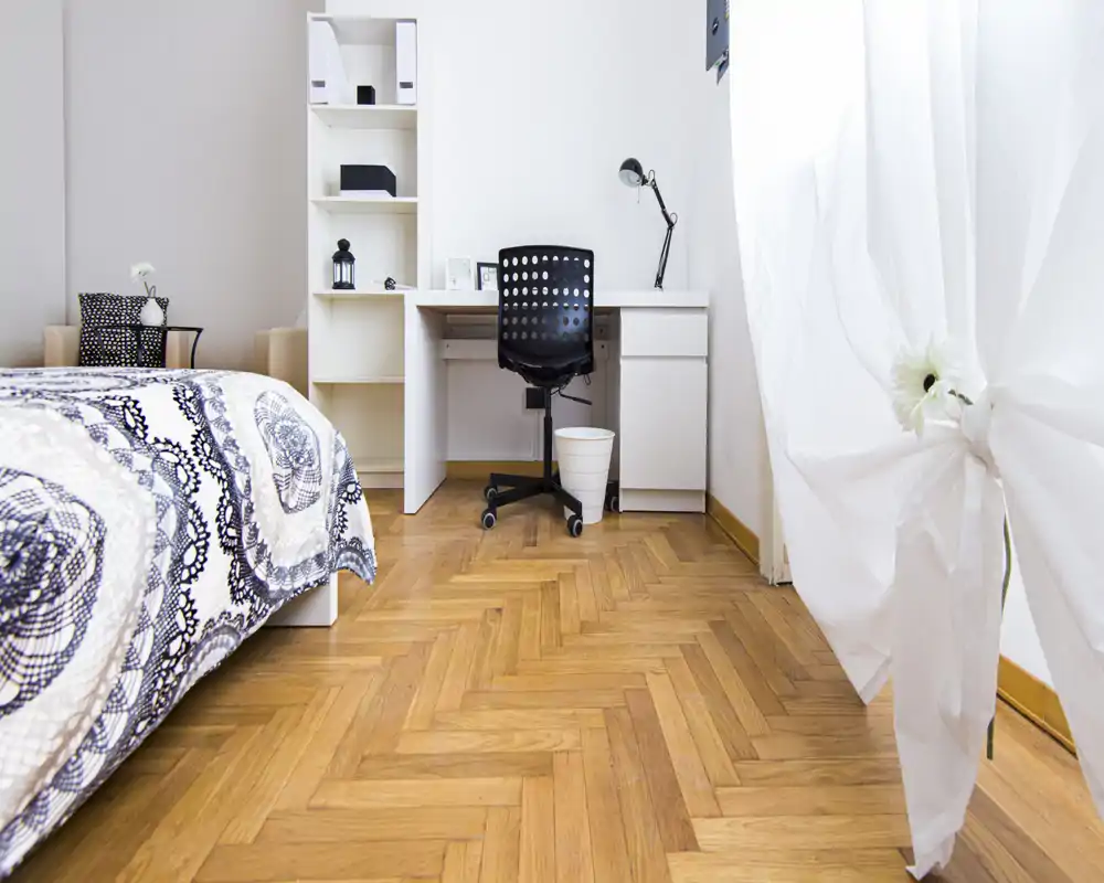 Bright, tidy bedroom with a patterned bedspread, a white study desk and shelving, and natural light from a large window—ideal for a calm, minimalist stay.
