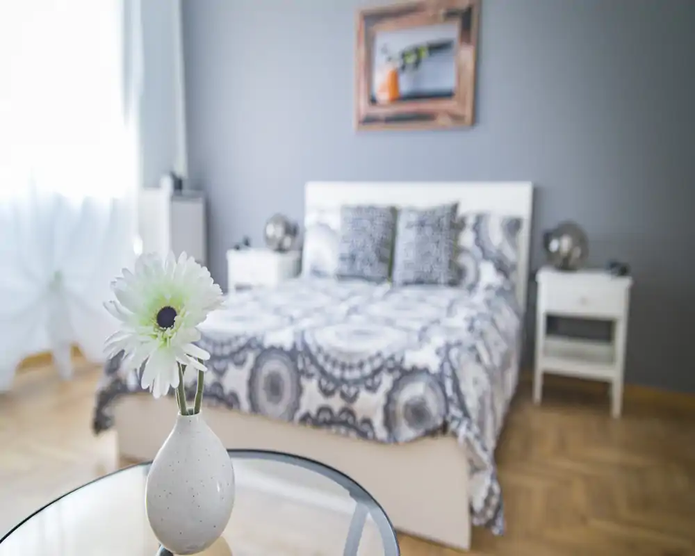 Bright, styled bedroom with patterned bedding, two bedside tables and soft natural light; foreground decorative flower adds depth.