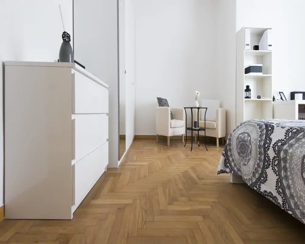 Bright, modern bedroom corner with patterned bedding, a white dresser, and a small sitting area. Natural light and warm parquet flooring create an inviting atmosphere.