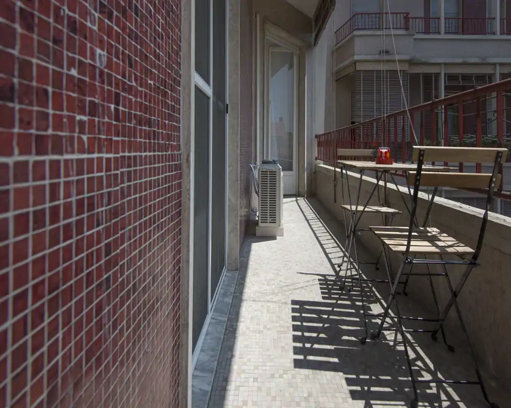 Balcone soleggiato e stretto con pavimento piastrellato, piccolo tavolo bistrò e sedie pieghevoli — un angolo esterno intimo con vista sulla città e ottima luce naturale.