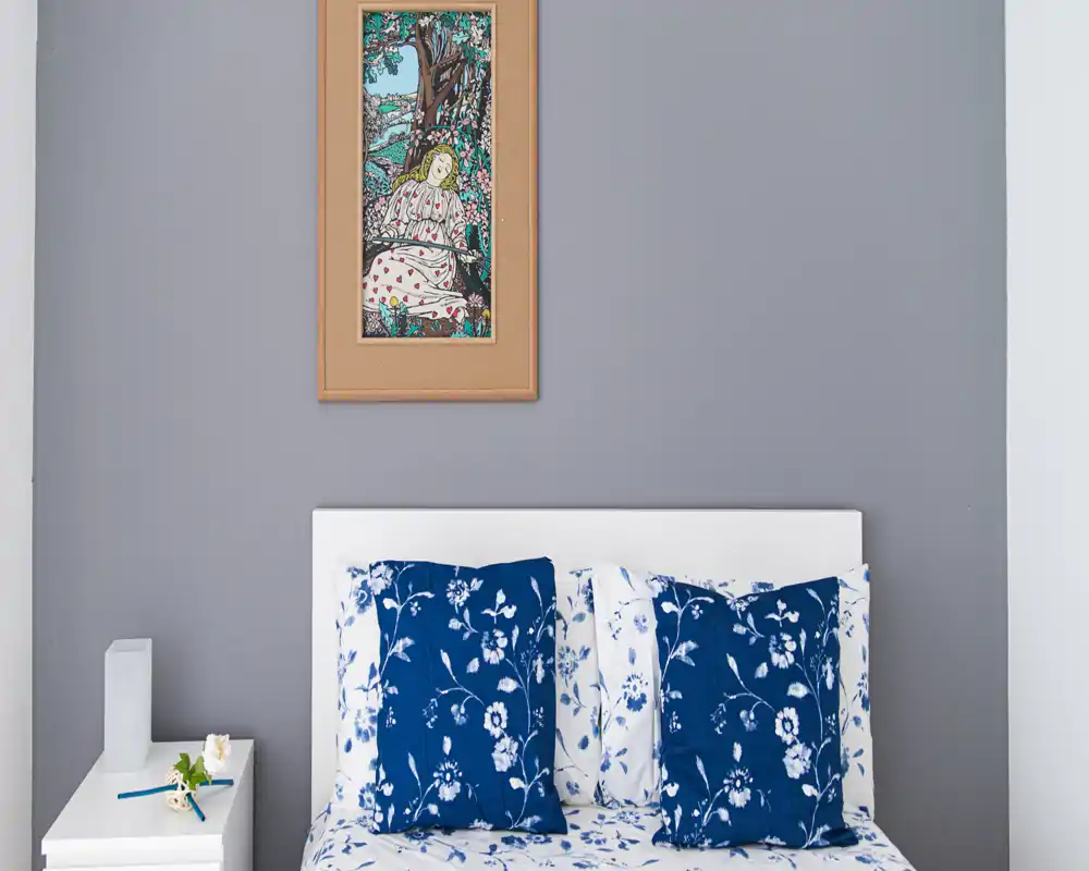 Camera da letto luminosa e ordinata con testiera bianca e cuscini floreali blu; un quadro elegante e un piccolo comodino completano l’atmosfera accogliente.