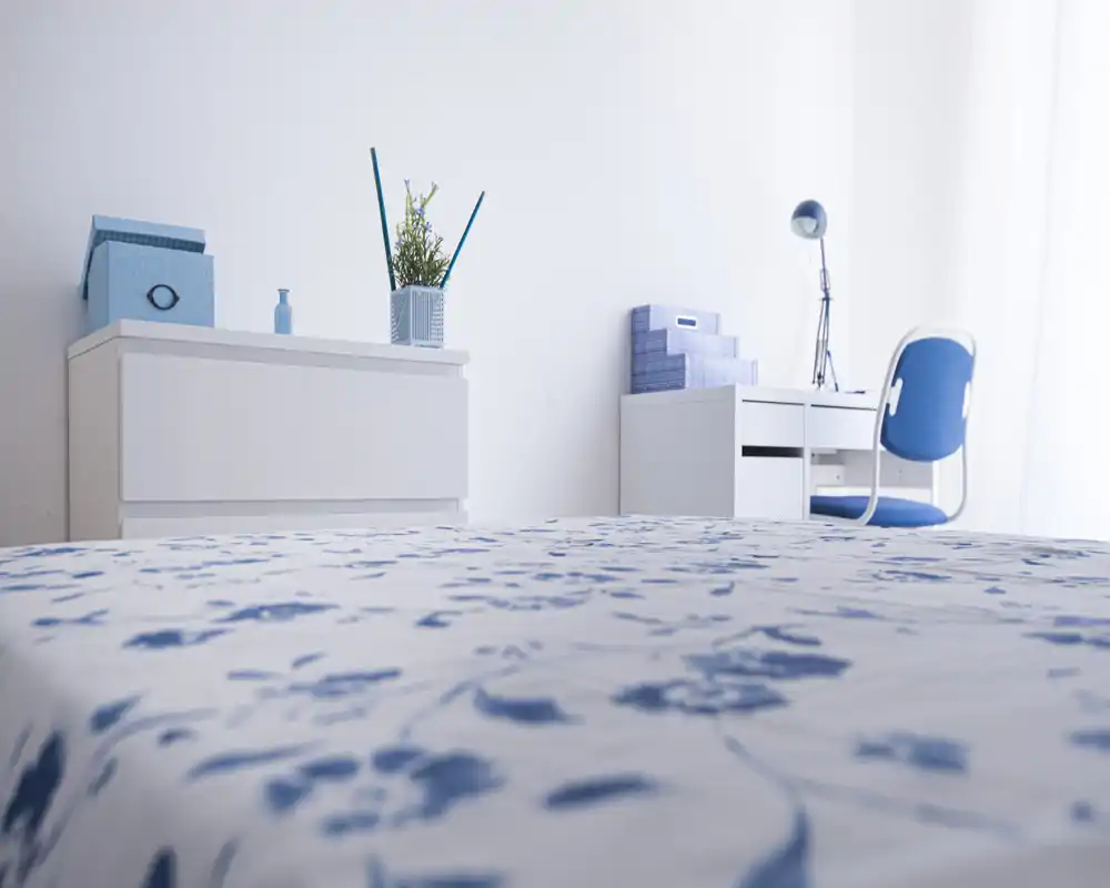 Camera da letto luminosa e minimal con un letto a fantasia blu e bianca in primo piano, un comò bianco e una scrivania con sedia blu vicino alla finestra. Tessuti leggeri e scatole coordinati creano un ambiente ordinato e arioso, ideale per annunci.