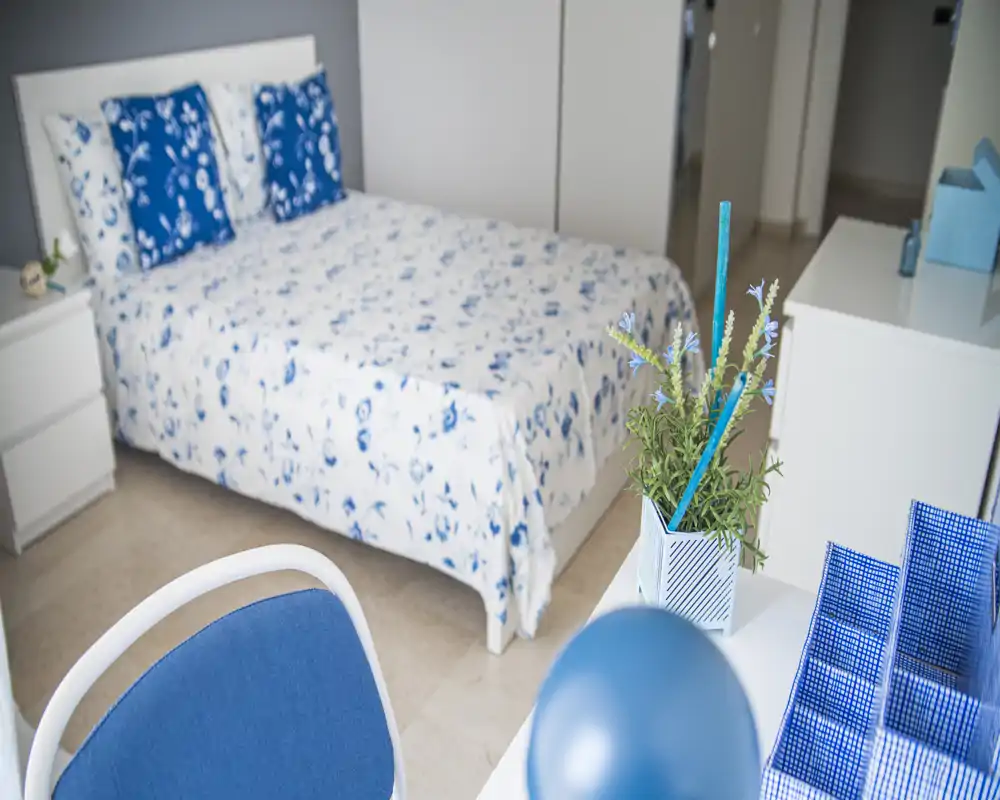 Camera da letto luminosa e ben arredata con copriletto floreale blu e bianco, cuscini coordinati e mobili bianchi semplici; luce naturale gradevole e dettagli decorativi curati.