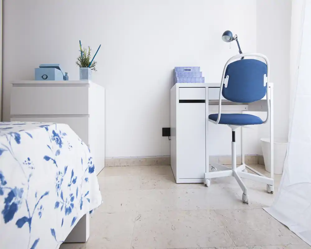 Angolo camera luminoso e minimal con copriletto bianco e blu a motivo, un piccolo scrittoio bianco e una sedia da ufficio blu — perfetto per una stanza compatta e ariosa.