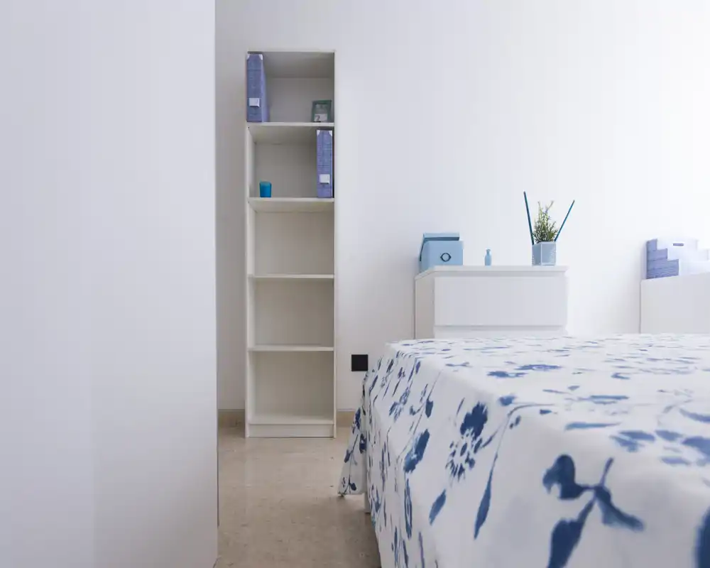 Camera da letto minimalista con copriletto bianco e blu, mensole bianche e comodino, illuminazione naturale soffusa — luminosa, ordinata e adatta a un annuncio di affitto.
