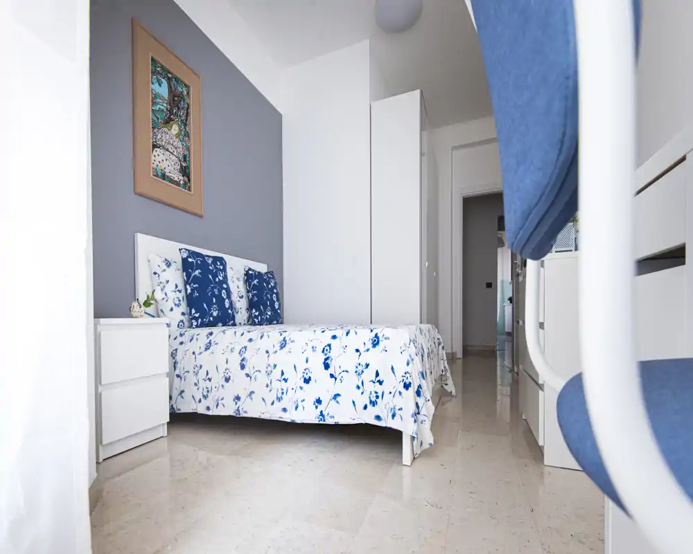 Camera da letto luminosa e ordinata con letto matrimoniale coperto da biancheria floreale blu e bianca, cuscini coordinati e mobili bianchi minimal. Parete grigio tenue, quadretto decorativo e luce naturale abbondante.