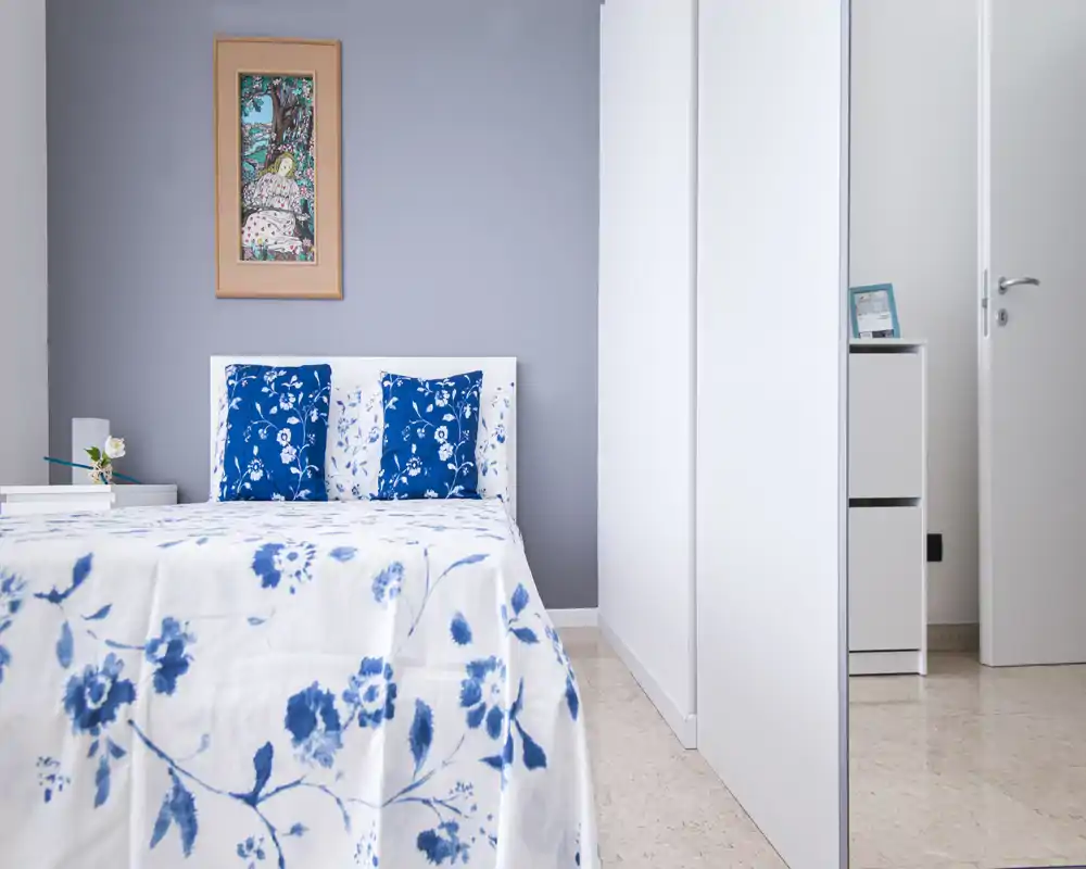 Camera da letto luminosa e minimale con testiera bianca e biancheria a fiori blu. La stanza è ordinata, ben illuminata e arredata con cuscini coordinati e una stampa incorniciata sopra il letto.