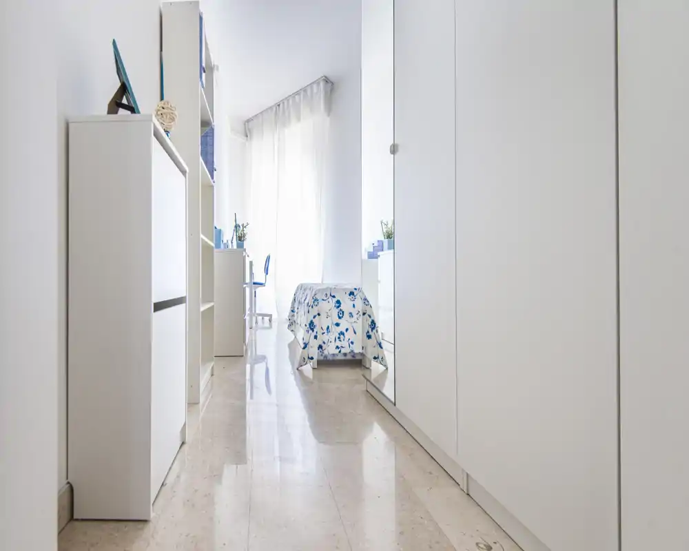 Camera da letto singola luminosa e minimal con copriletto floreale, armadi a muro e una piccola scrivania vicino alla finestra con tende leggere che filtrano la luce naturale.