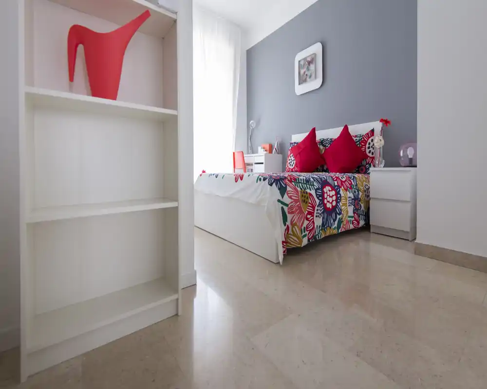 Bright, modern bedroom with a colorful bedspread and red accent cushions; natural light from a window highlights clean lines and minimalist furniture.
