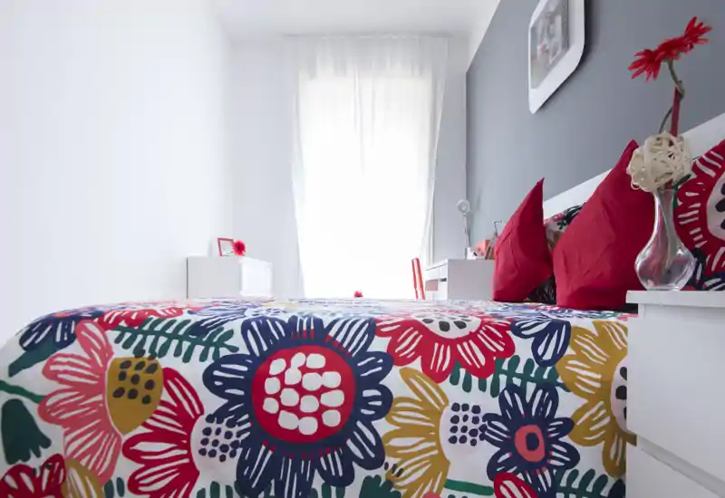 Dormitorio luminoso y moderno con colcha floral colorida, cojines rojos como acento y luz natural procedente de una gran ventana. Muebles blancos y una pared gris para un aspecto fresco y contemporáneo.