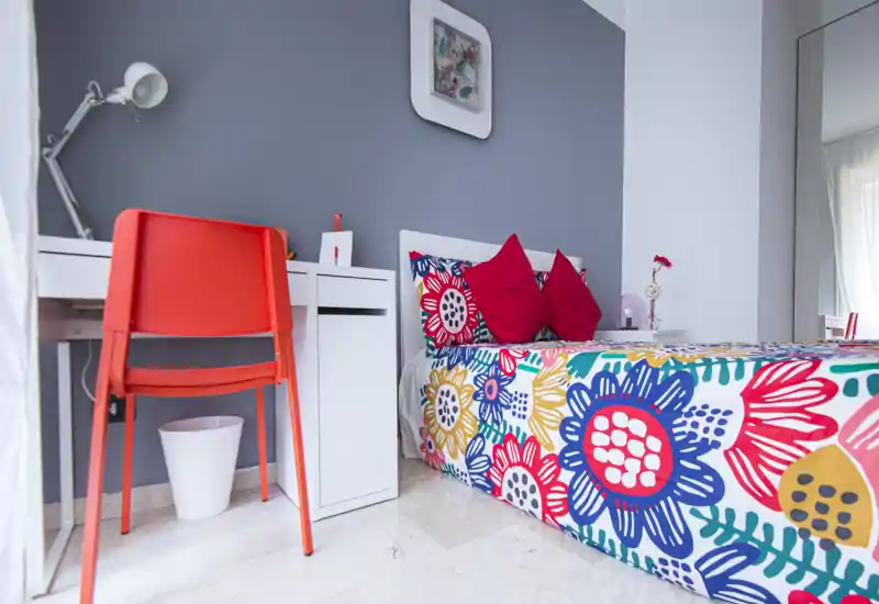 Dormitorio individual luminoso y colorido con funda nórdica floral, dos cojines rojos y un escritorio blanco con silla naranja — perfecto para un alquiler vibrante y bien iluminado.