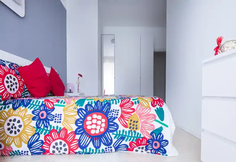 Dormitorio moderno y luminoso con colcha floral colorida y cojines rojos. Paredes blancas y un armario alto crean un ambiente fresco y minimalista.