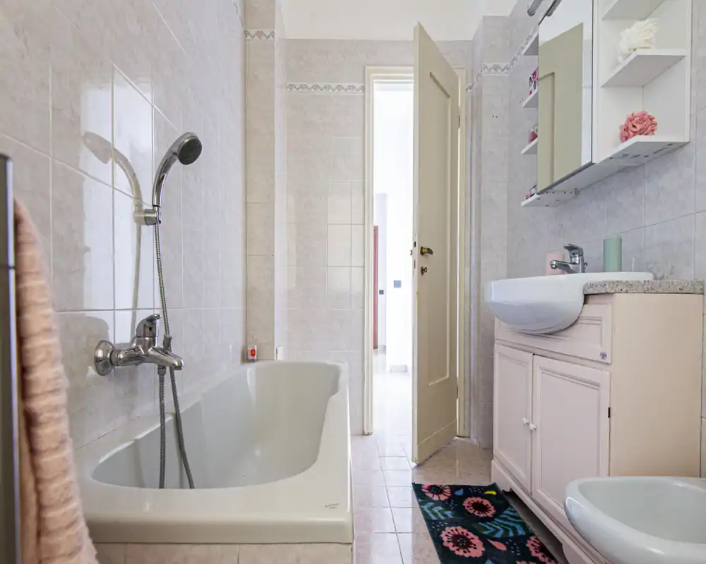 Salle de bains propre et lumineuse avec baignoire, douchette, lavabo avec meuble et miroir; carrelage clair et tapis coloré apportant une touche chaleureuse.