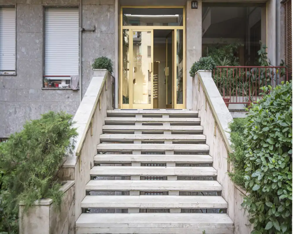 Entrada simétrica de piedra con amplias escaleras exteriores que conducen a una puerta de cristal, flanqueada por vegetación y macetas: ideal para mostrar el acceso y el atractivo exterior del edificio.