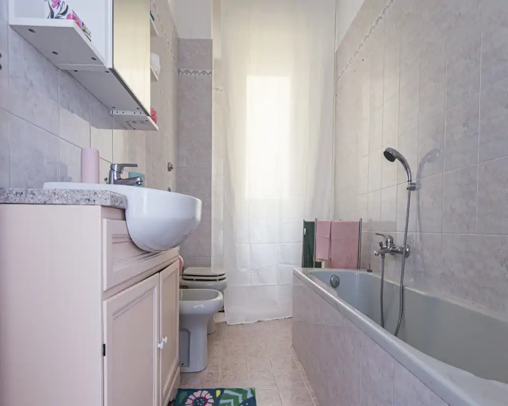 Baño limpio y bien iluminado con bañera, teleducha, lavabo con mueble y bidé/inodoro visibles; azulejos rosa suave y una cortina traslúcida que aportan una atmósfera ligera y agradable.