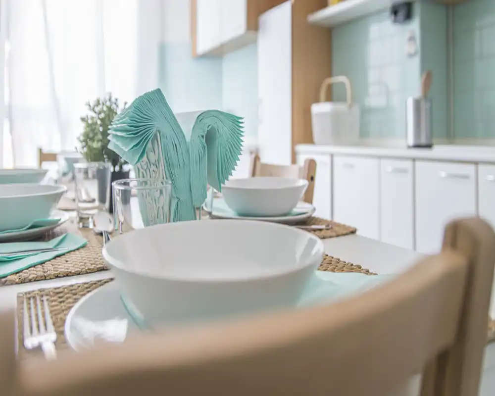Zona de cocina/comedor luminosa y cuidada con primer plano de una mesa puesta: cuencos blancos, individuales de fibra y servilletas turquesa plegadas, iluminada por luz natural.