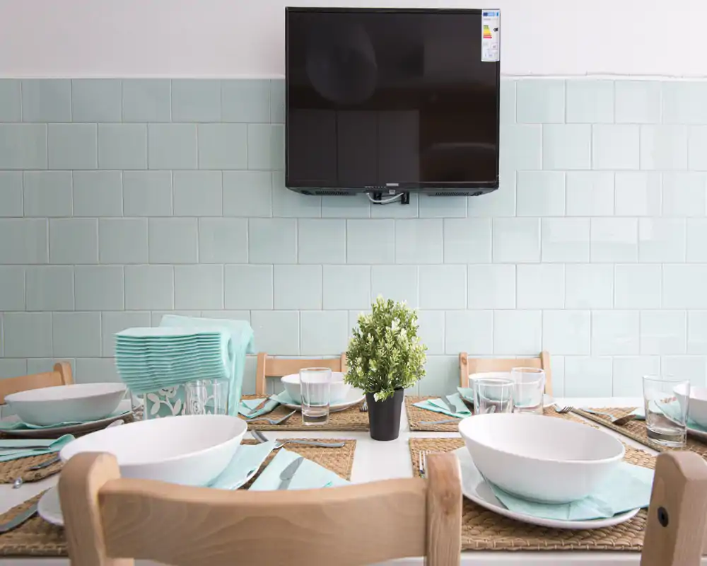 Zona de comedor decorada con mesa puesta: salvamanteles de fibras, boles blancos y servilletas turquesa alrededor de una planta central y una TV en la pared — luminosa, ordenada y lista para invitados.