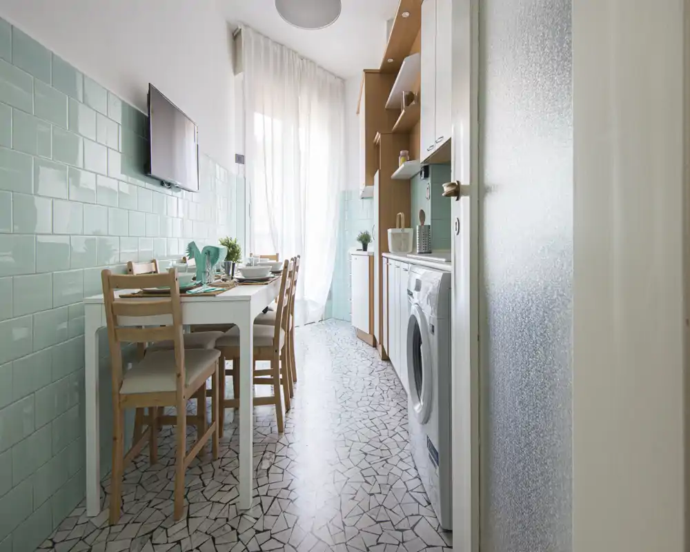 Cocina estrecha y luminosa con azulejos verde menta, mesa para cuatro y electrodomésticos integrados, incluida la lavadora. Cortinas traslúcidas suavizan la luz y las sillas de madera aportan calidez.