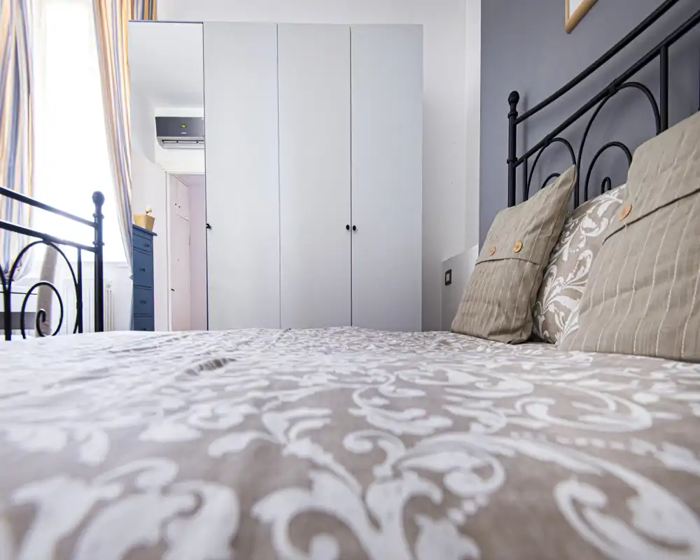 Bright, cozy bedroom with decorative pillows and a patterned duvet; large wardrobe and window provide natural light and storage.