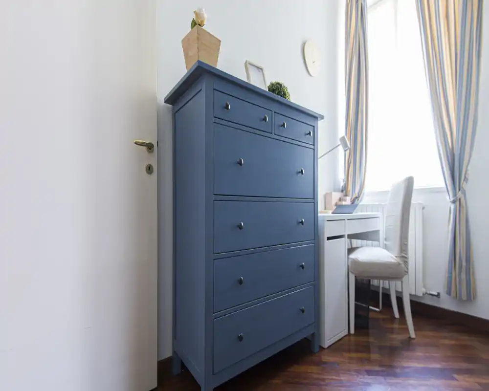 Bright, well-styled bedroom corner with a tall blue chest of drawers, a compact white desk and a cushioned chair by a sunny window with striped curtains.