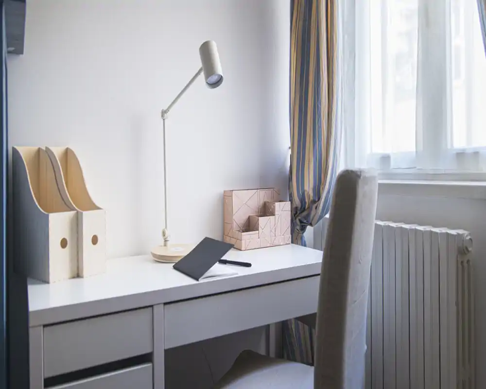 Bright, minimalist workspace with a white desk, task lamp, organizer and upholstered chair beside a window with striped curtains — ideal for focused work or study.