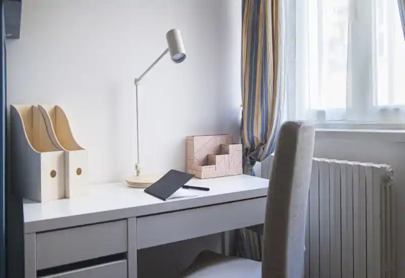 A bright, minimal home-office corner with a white desk, desk lamp, upholstered chair and window with striped curtains — tidy and well-lit for work or study.